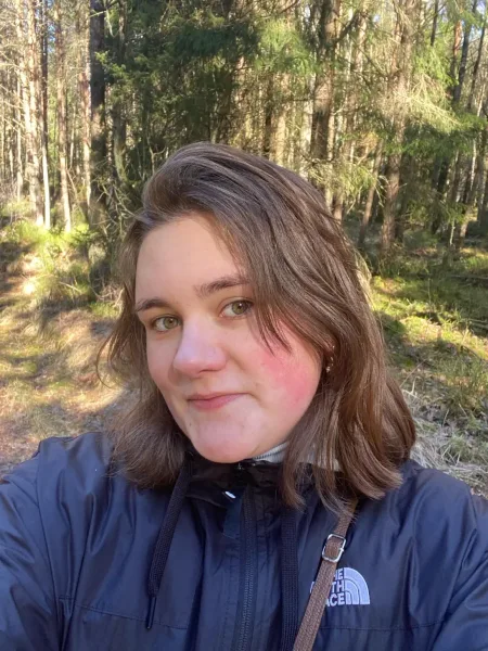 Mitarbeiter_Foto_Janne Eine Frau mit braunen Haaren und einer dunkelblauen Outdoor-Jacke, die Mitarbeiterin des Cafés "Mimi's Welt - Das Kindercafé" ist und lächelt. Im Hintergrund ist ein Wald zu sehen.