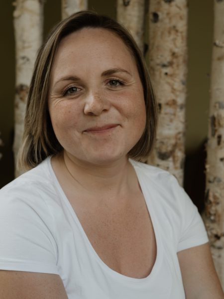 Mitarbeiter_Foto_Sophie Eine Frau mit braunen Haaren und einem weißen T-Shirt, die Mitarbeiterin des Cafés "Mimi's Welt - Das Kindercafé" ist und lächelt. Im Hintergrund sind echte Birkenstämme zu sehen